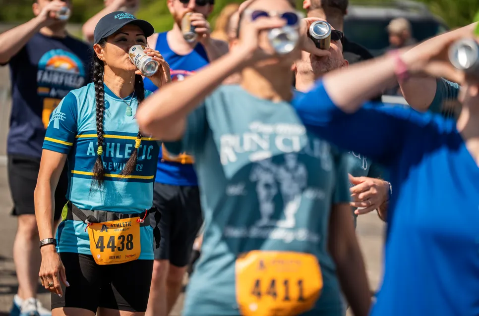 Runners in athletic wear, some holding cans, with race bibs visible, suggesting a post-race celebration or a unique race event.