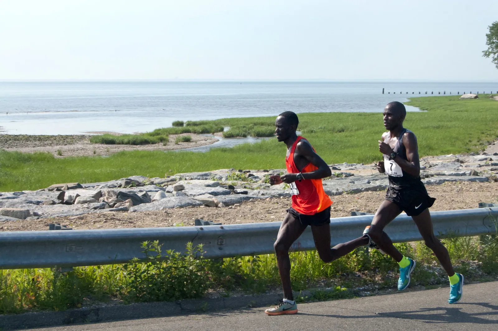 Two male runners race along a road with a waterfront view.