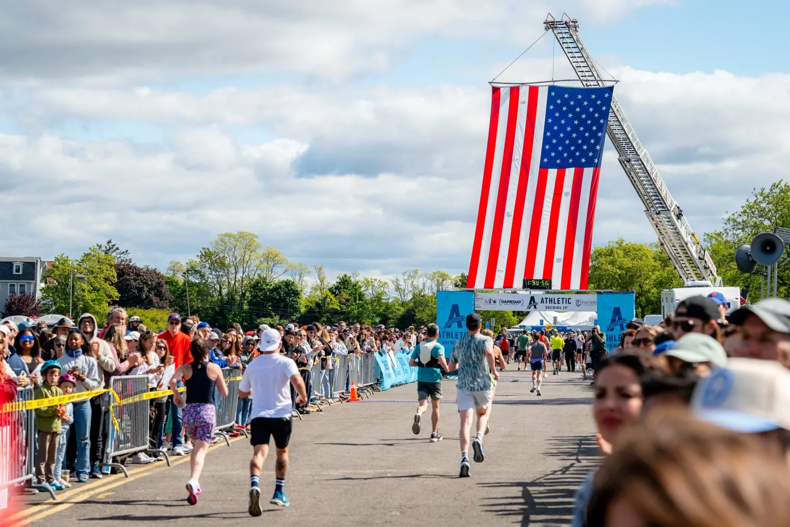 Runners cross the finish line of a road race, cheered on by a large crowd, with a large American flag hanging from a fire truck ladder overhead.