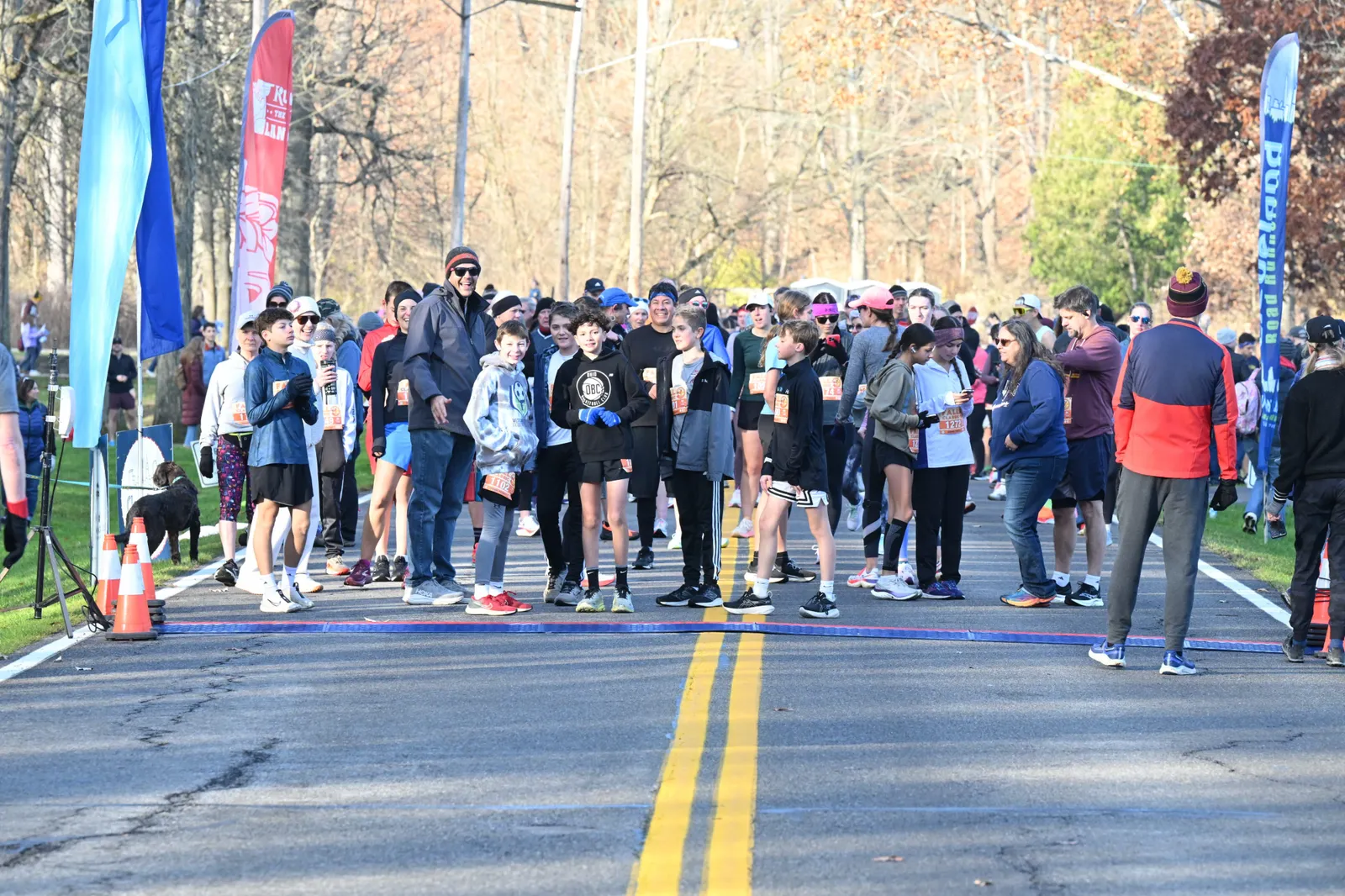A diverse crowd of runners, including children and adults, gathered at the start line of a road race on a sunny day, with colorful flags and trees in the background.
