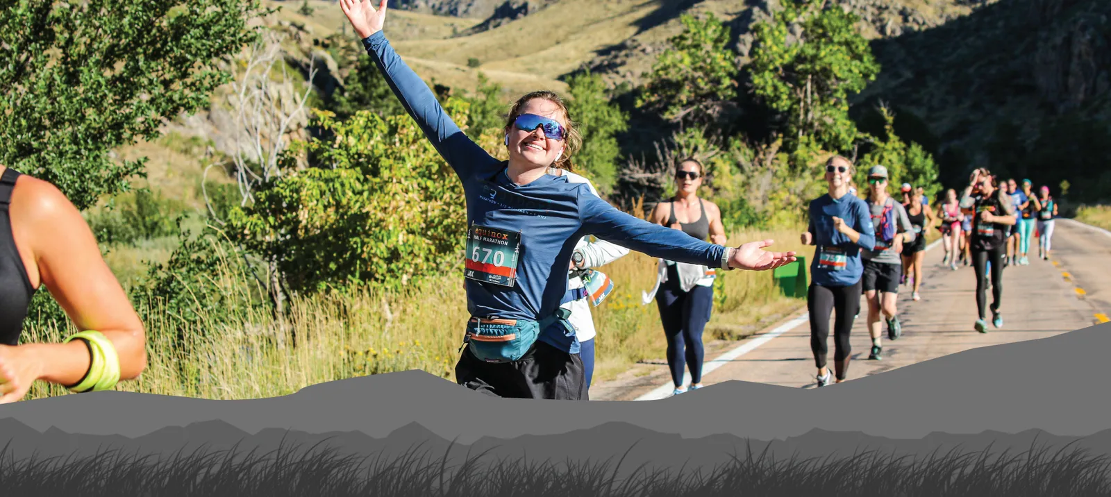 A smiling female runner with her arms raised in celebration, running on a road with other participants in the background, set against a scenic, hilly landscape.
