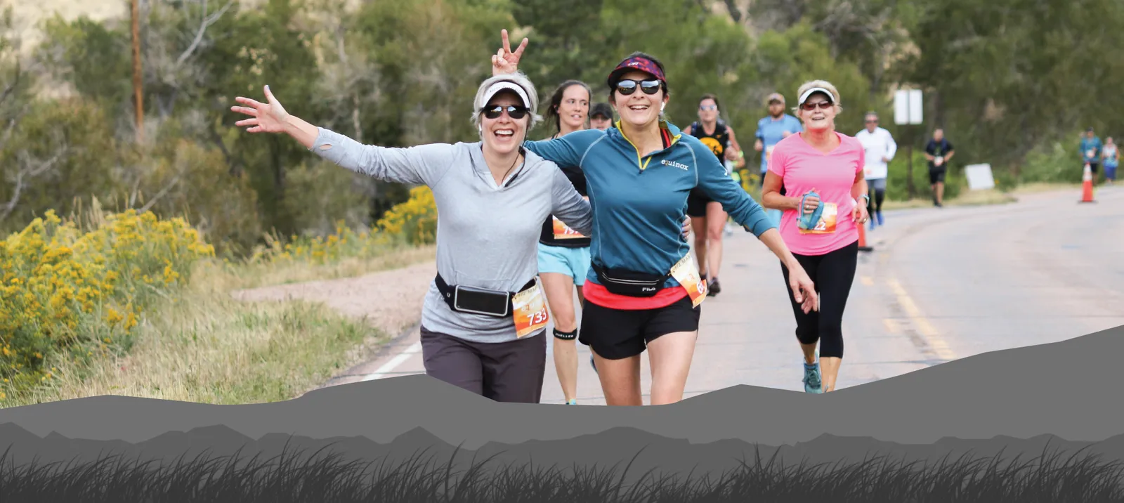 Two women smiling and running, one giving a peace sign, with other runners in the background on a paved road.