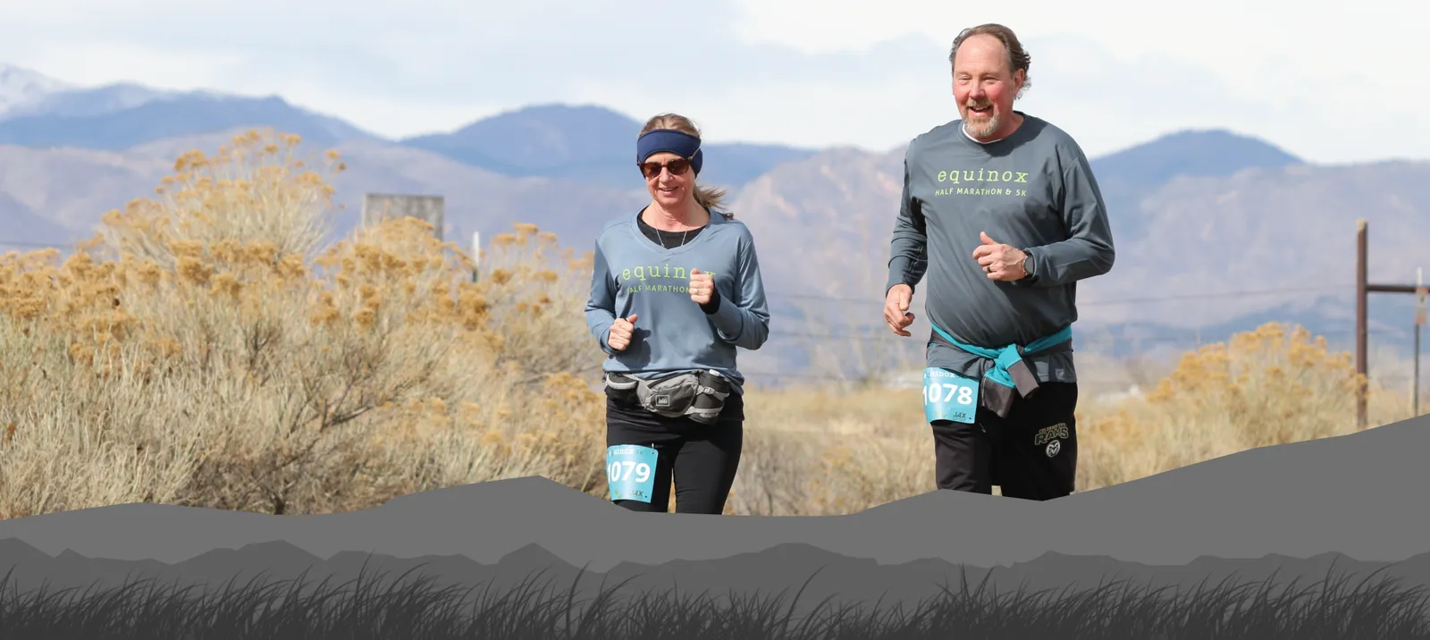 Two runners, a man and a woman, smiling as they run on a road with mountains and dry brush in the background.