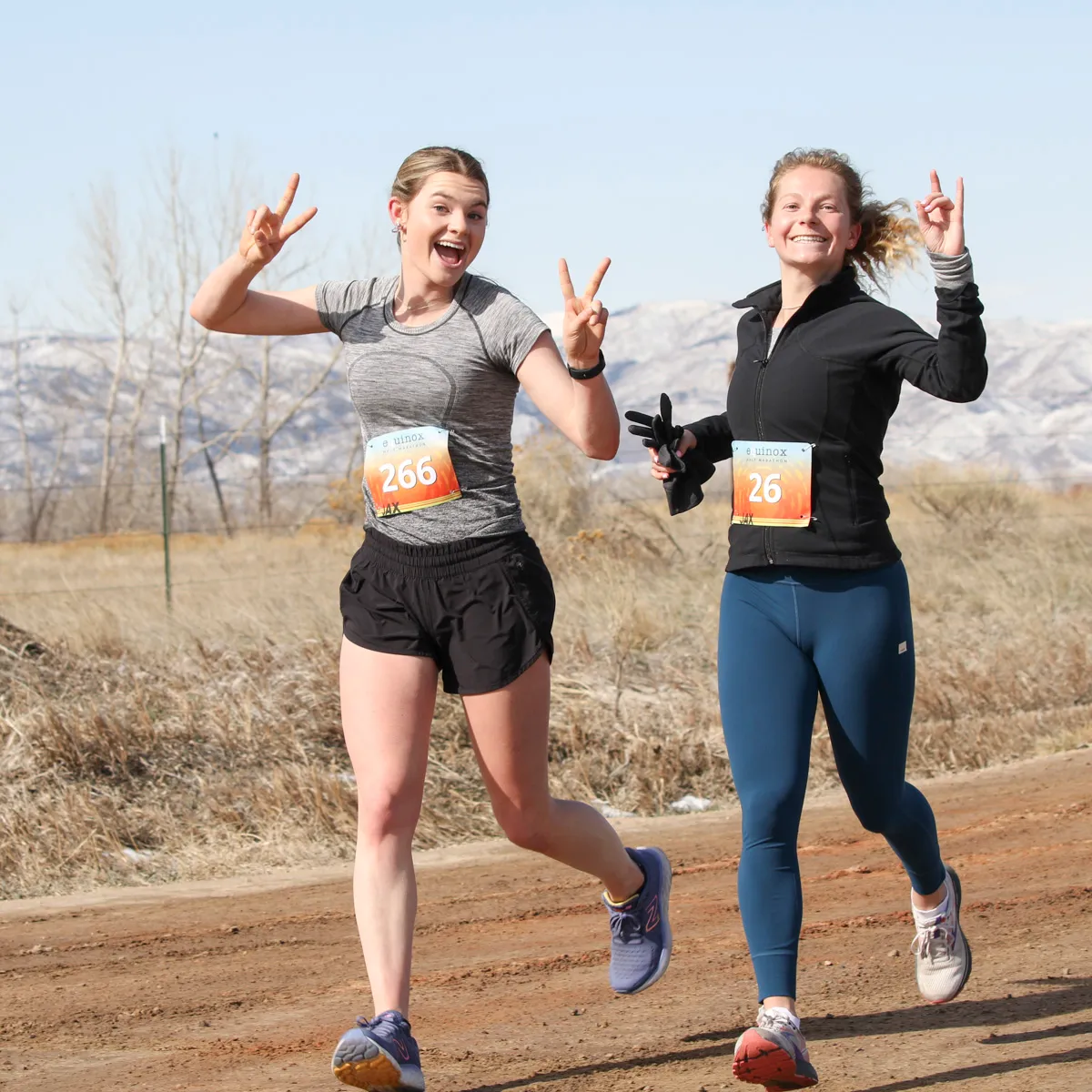 Two female runners smiling and giving peace signs while running on a dirt road with mountains in the background.