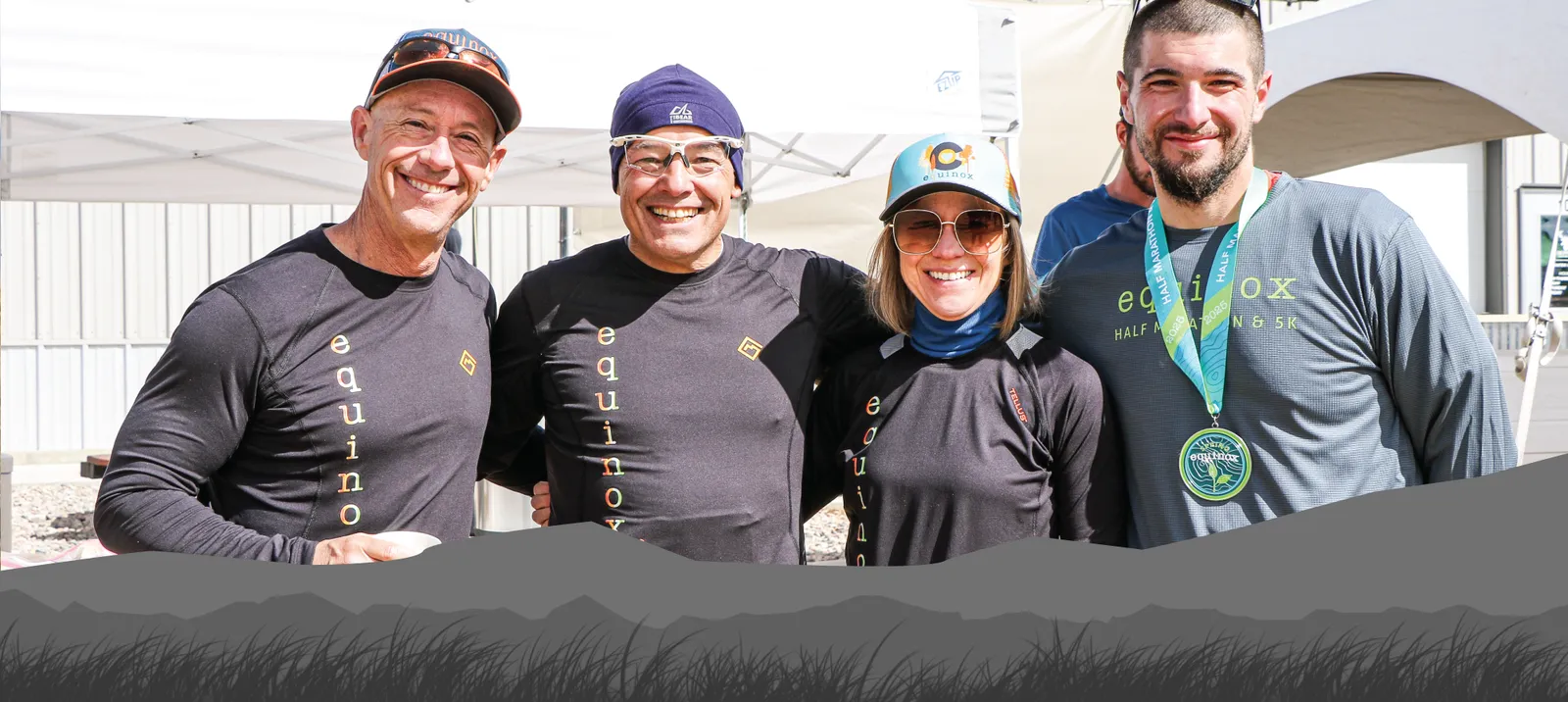 Four smiling people, two men and two women, pose for a photo. One man wears a race medal around his neck, and the others wear race-related apparel. They appear to be at an outdoor event.