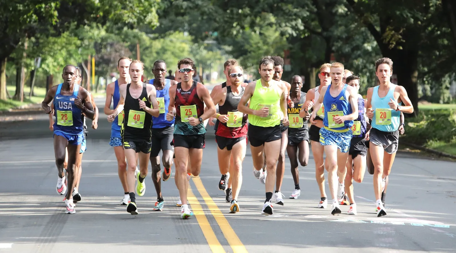 A group of male runners in a race, with bib numbers visible, running on a road with trees in the background.
