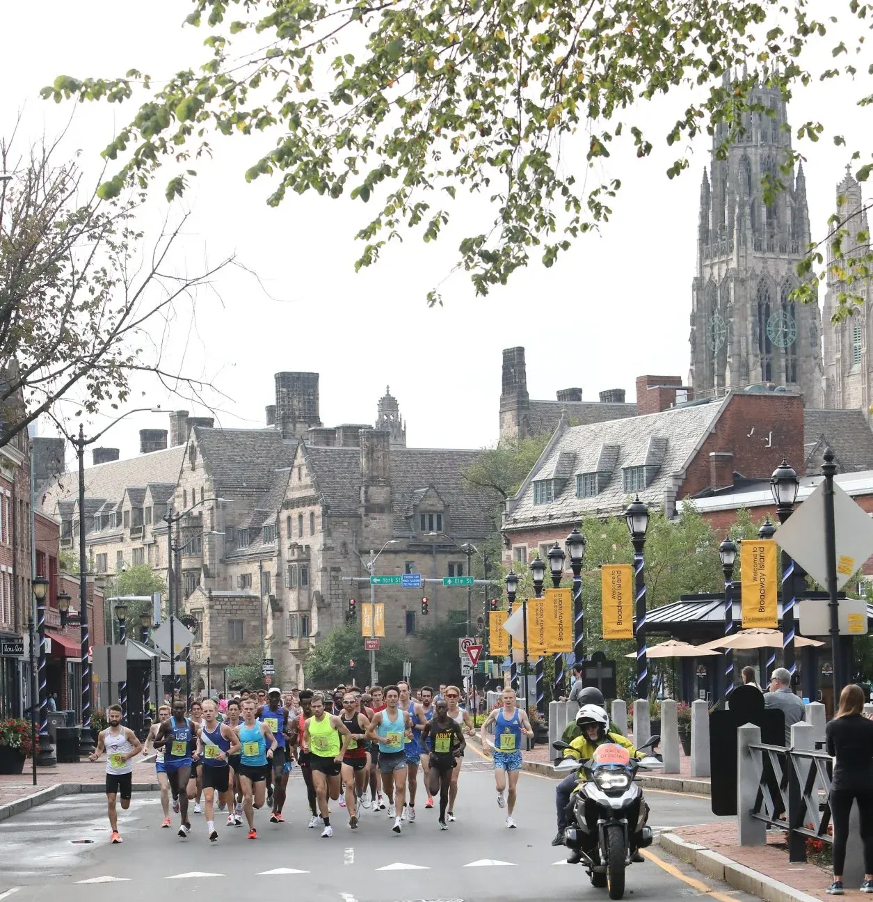 A group of runners race down a city street lined with historic buildings, with a motorcycle escorting them.