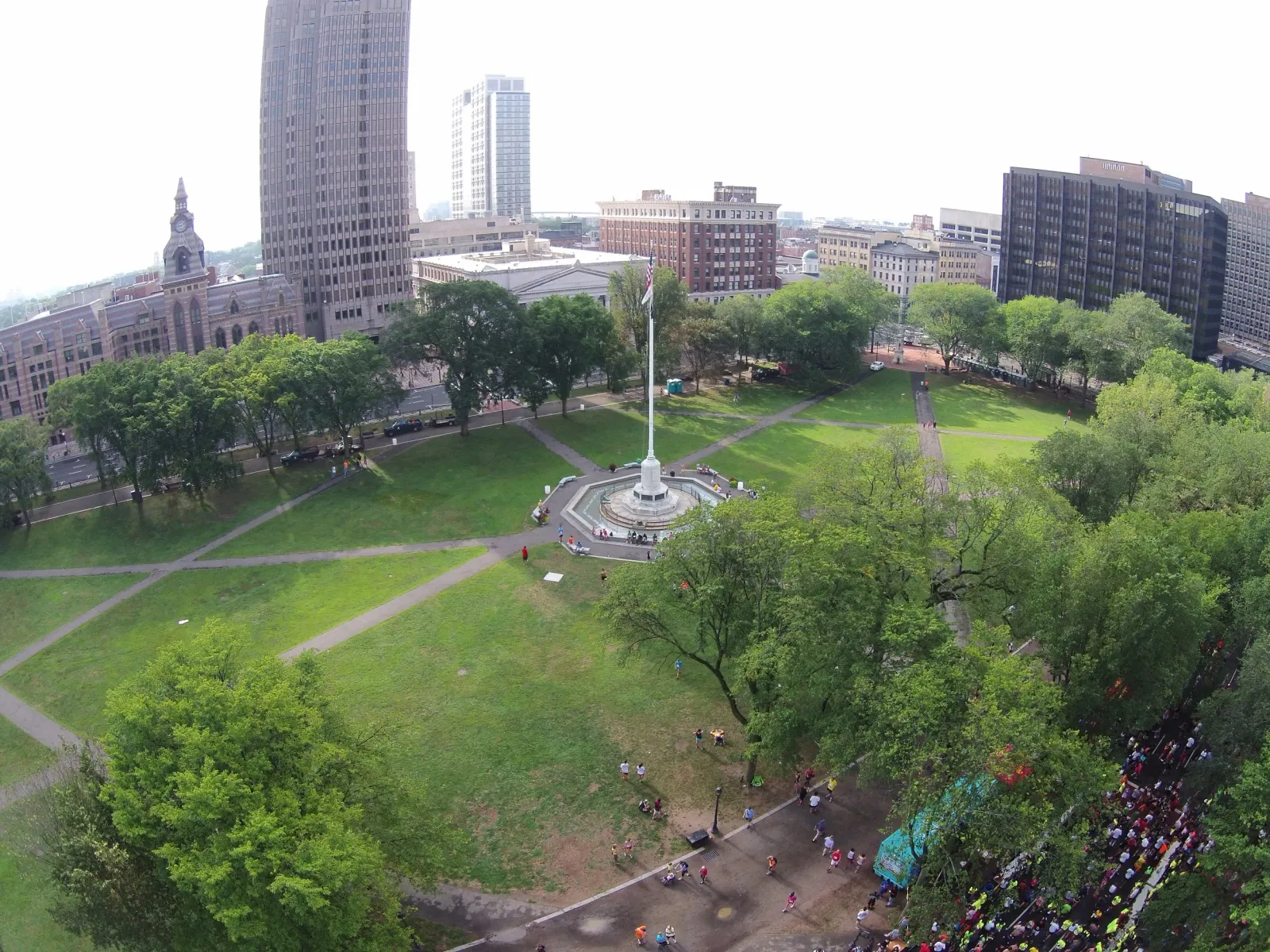 A high-angle view of a large crowd of runners on a road in an urban park, with tall buildings in the background.