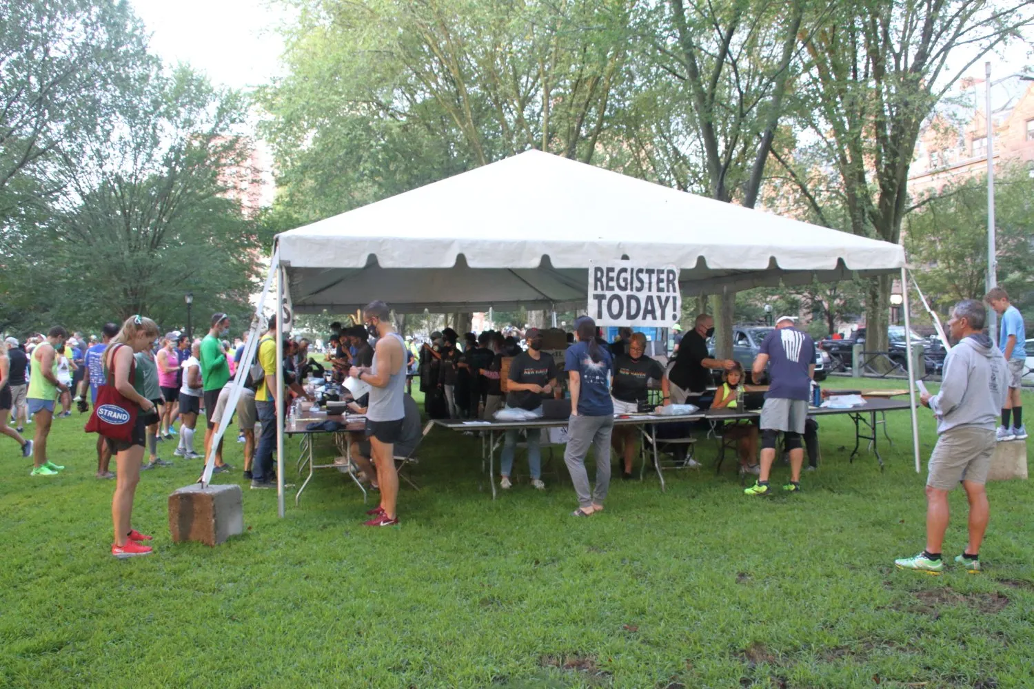 Runners gather under a tent with a 'REGISTER TODAY!' sign, indicating pre-race registration and activity.