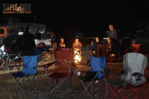 People gathered around a campfire at night, with chairs set up and a few vehicles in the background.