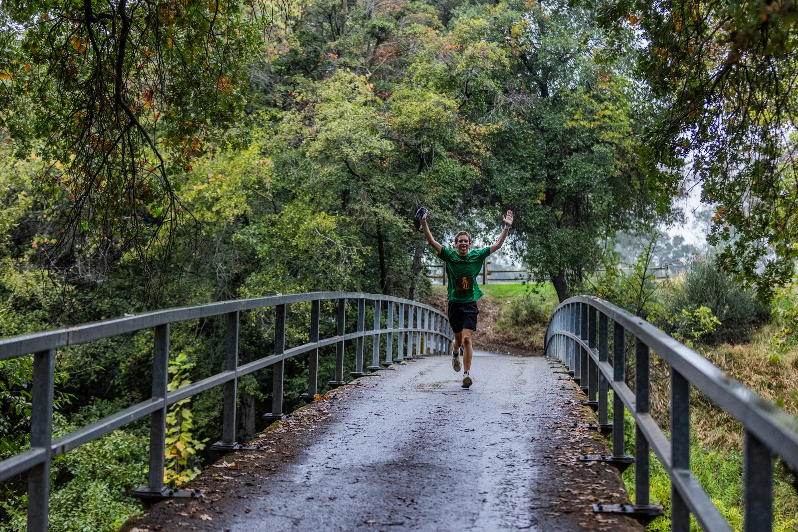 A runner with arms raised in celebration runs across a bridge on a wooded trail.