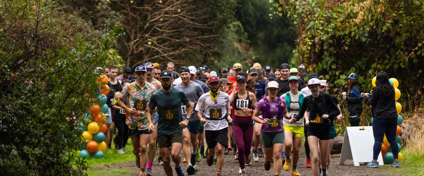 A large group of runners starting a trail race, with balloons and greenery in the background.