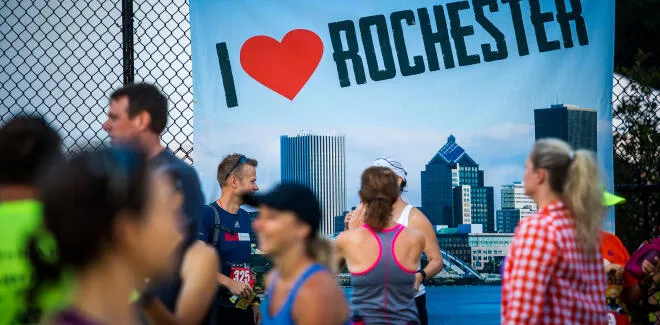 Runners gathered in front of a large banner that says 'I Love Rochester' with a cityscape in the background.