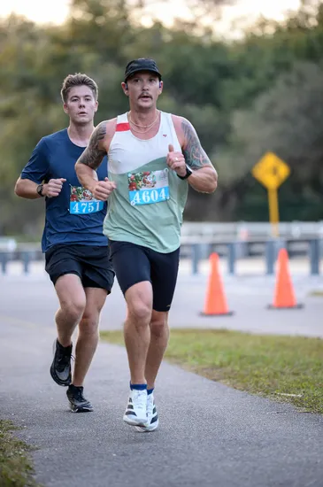 Two male runners on a paved path, one in front with bib number 4604 and a light green tank top, the other behind with bib number 4751 and a dark blue t-shirt.