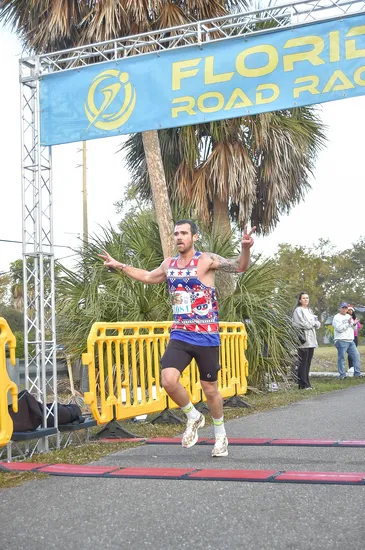 A male runner wearing a festive tank top crosses the finish line, holding up two fingers in a peace sign, with the 'Florida Road Race' banner overhead.