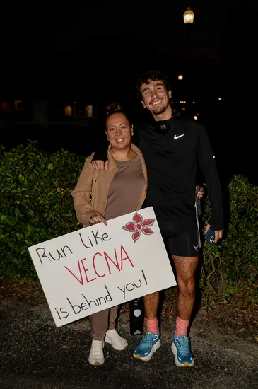 A woman and a man smile while holding a sign that says 'Run like VECNA is behind you!' with a drawing of a flower.