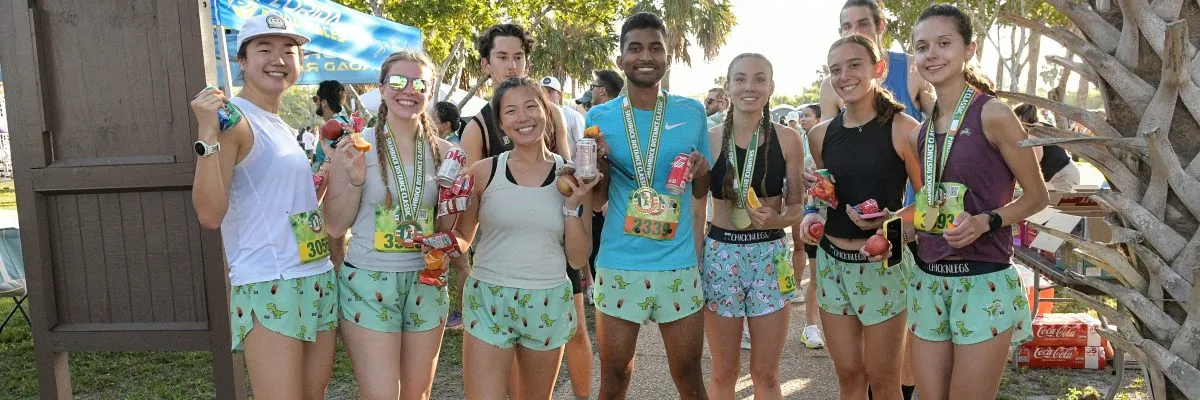 A group of smiling runners, some wearing medals, pose for a photo after a race, holding snacks and drinks.