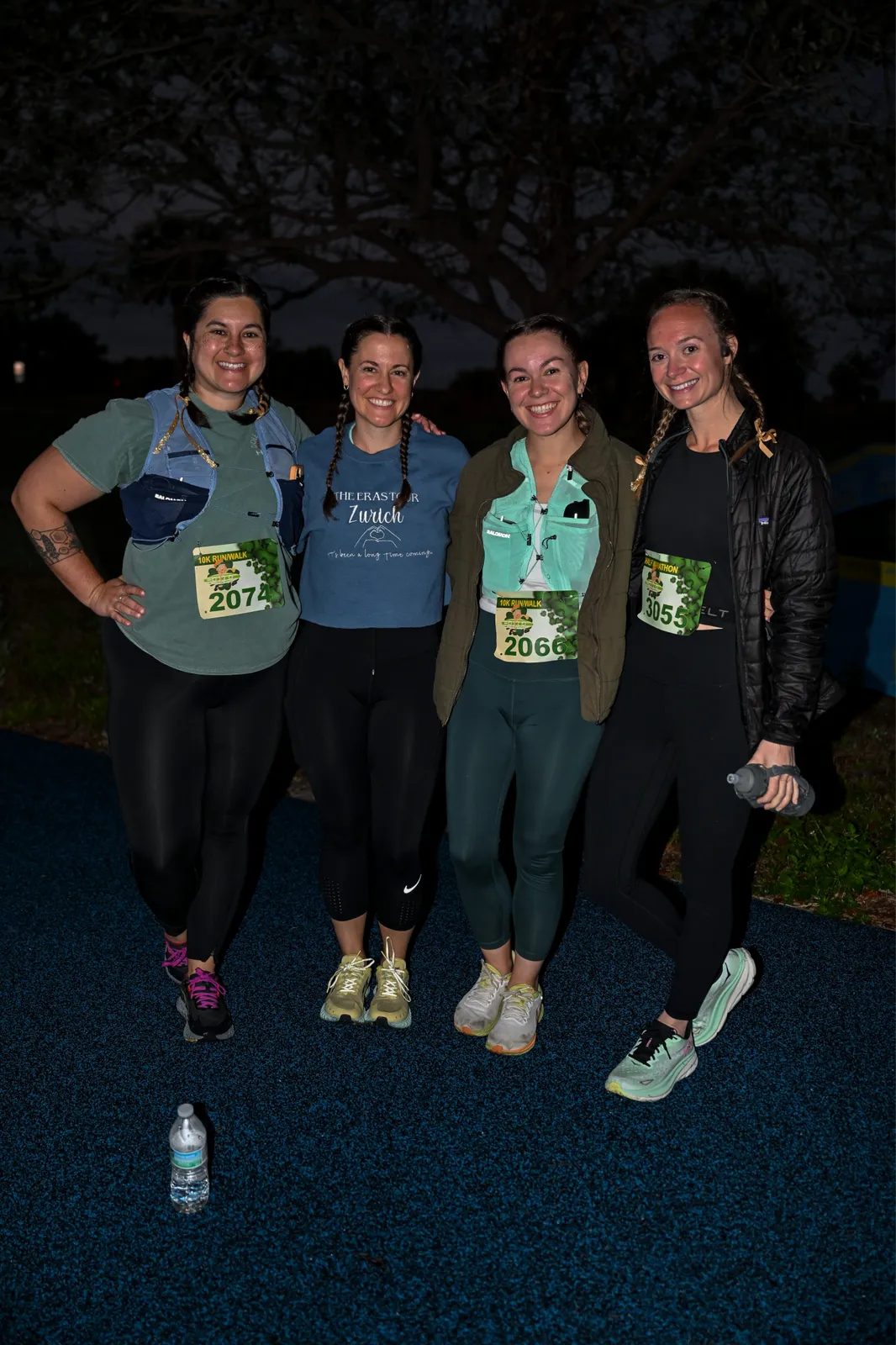 Four women smiling and posing for a photo, wearing race bibs and athletic clothing.