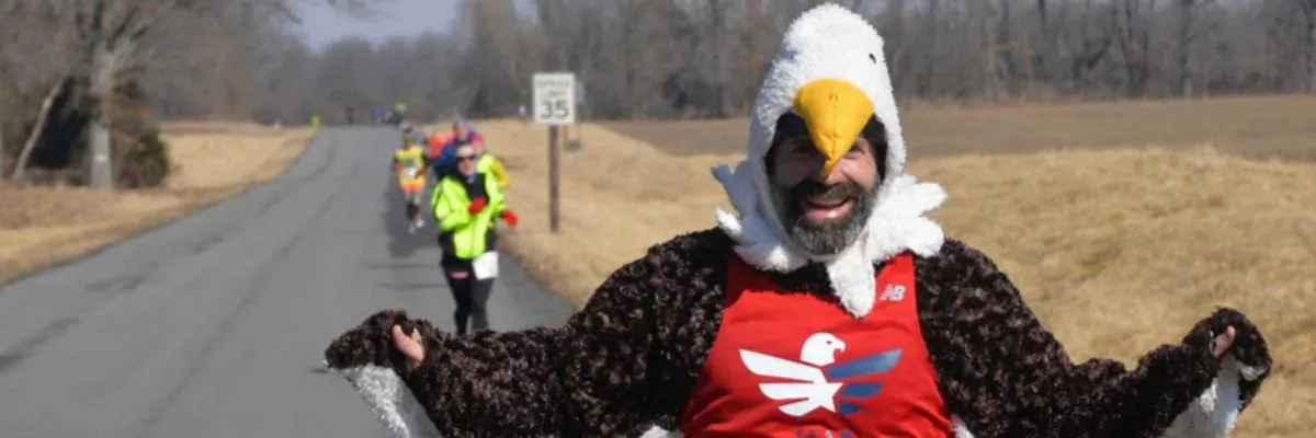 A runner in a full eagle costume with outstretched wings smiles at the camera, with other runners visible on a road in the background.