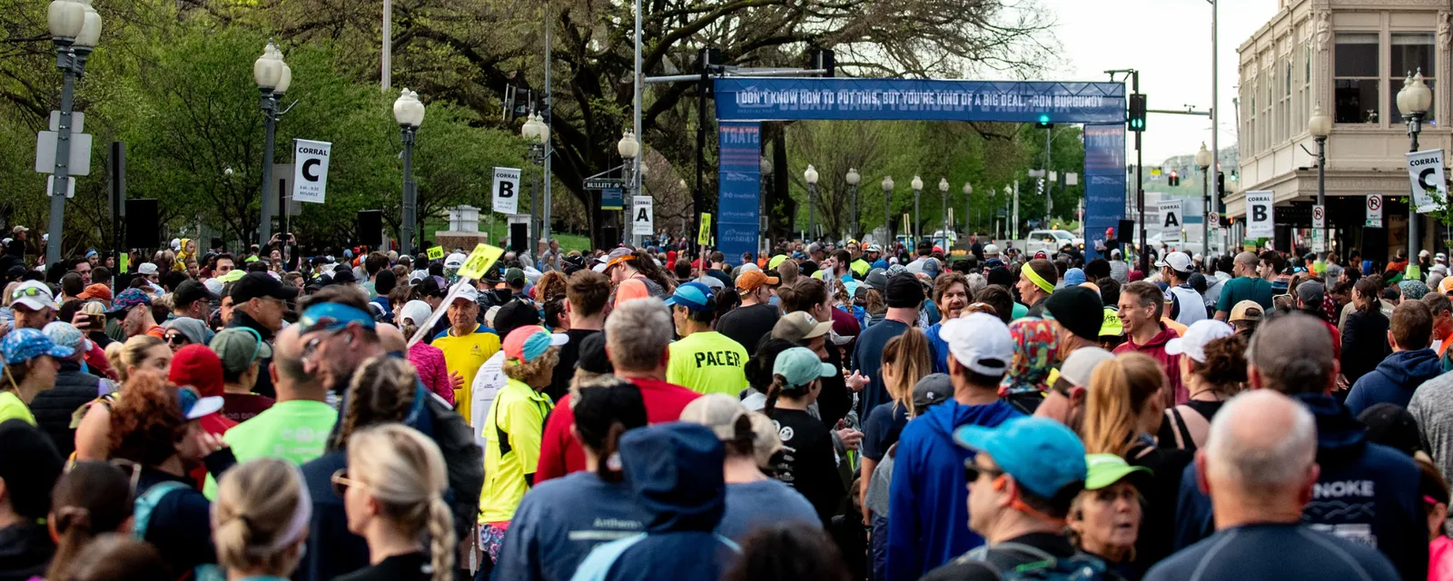 A large crowd of runners gathered at the start line of a race, with corrals marked 'A', 'B', and 'C' visible.