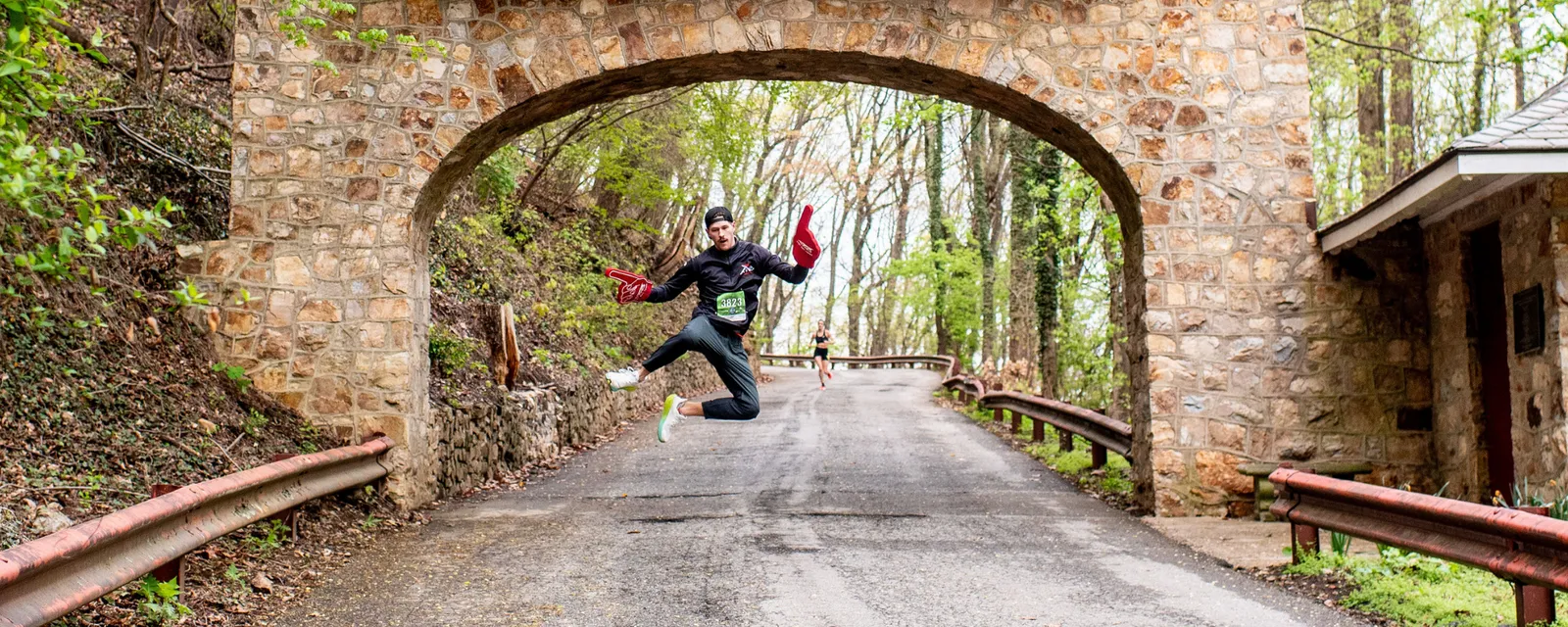A runner in mid-air jump under a stone archway on a road surrounded by trees, with another runner in the distance.