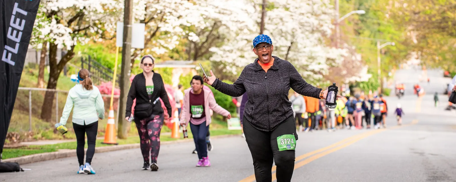 A joyful runner with arms outstretched and a wide smile, wearing bib number 3724, participates in a road race with other runners and spectators in the background.