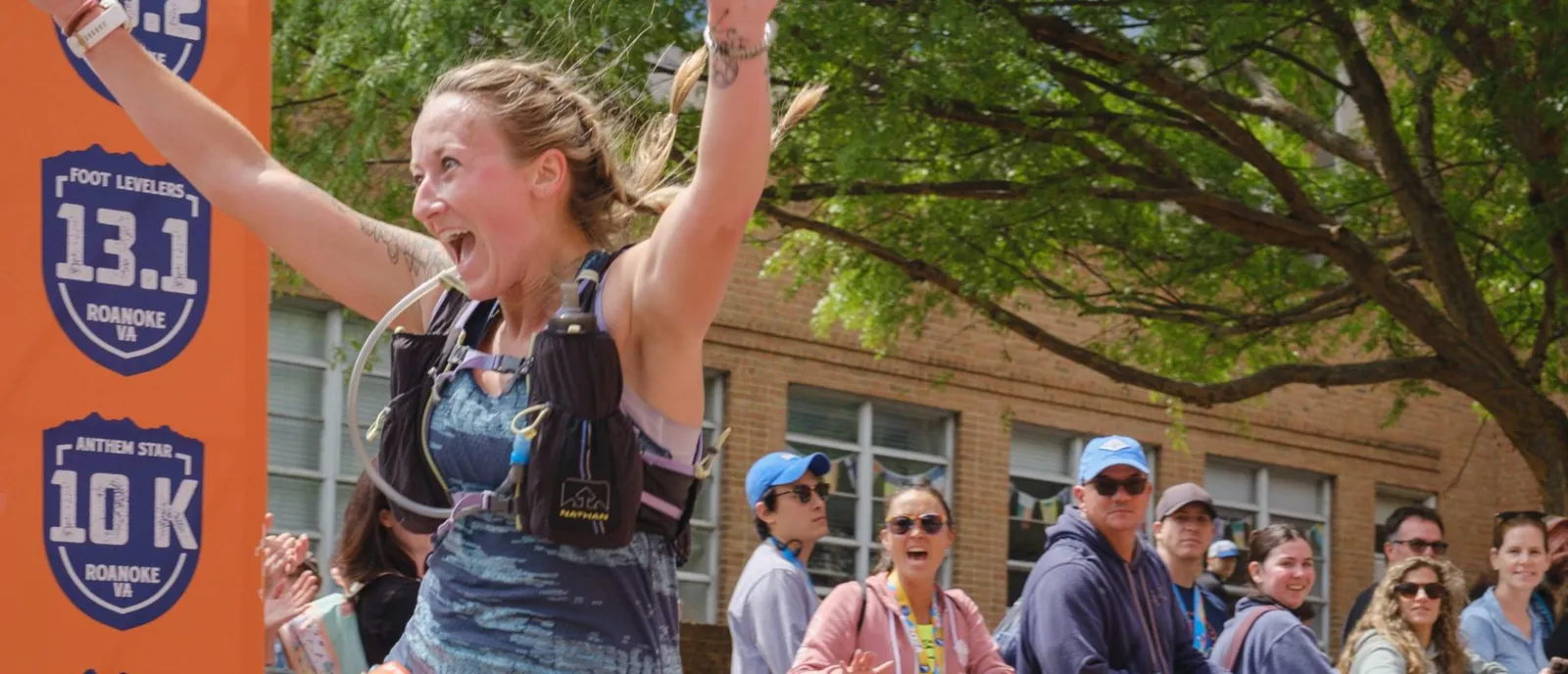 A female runner with her arms raised in triumph crosses the finish line, with a cheering crowd and race banners in the background.