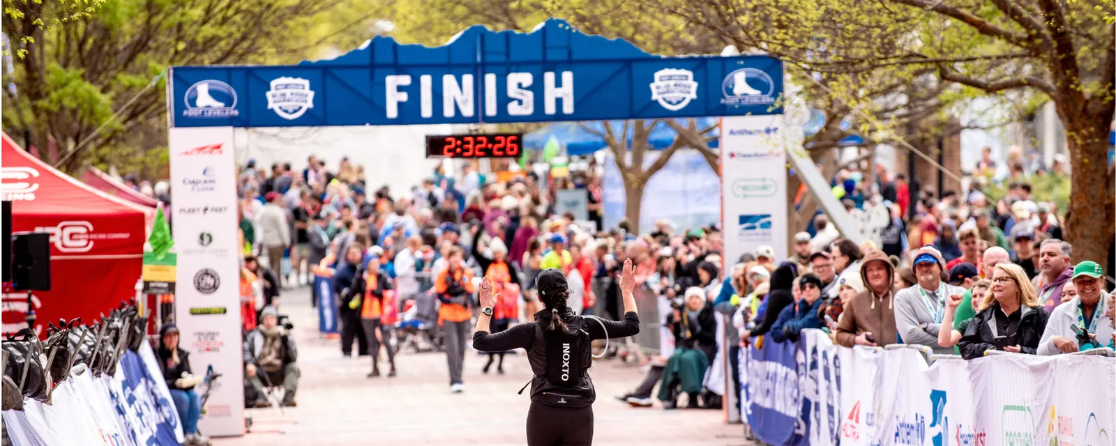 A runner gives a high-five as they approach the finish line at the Blue Ridge Marathon, with a large crowd watching and a digital clock displaying the race time.