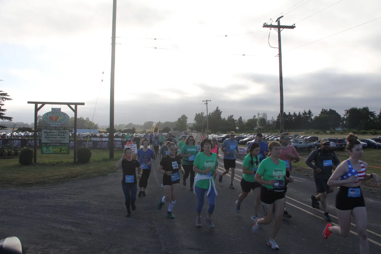 A group of runners are starting a race on a gravel road, with a sign for 'The Pumpkin Patch' visible in the background.
