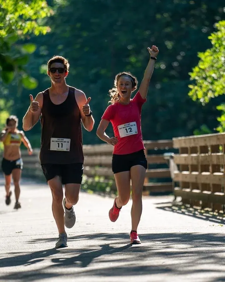 A male runner gives a thumbs up while a female runner celebrates with her arm in the air, both running on a paved path with trees in the background.