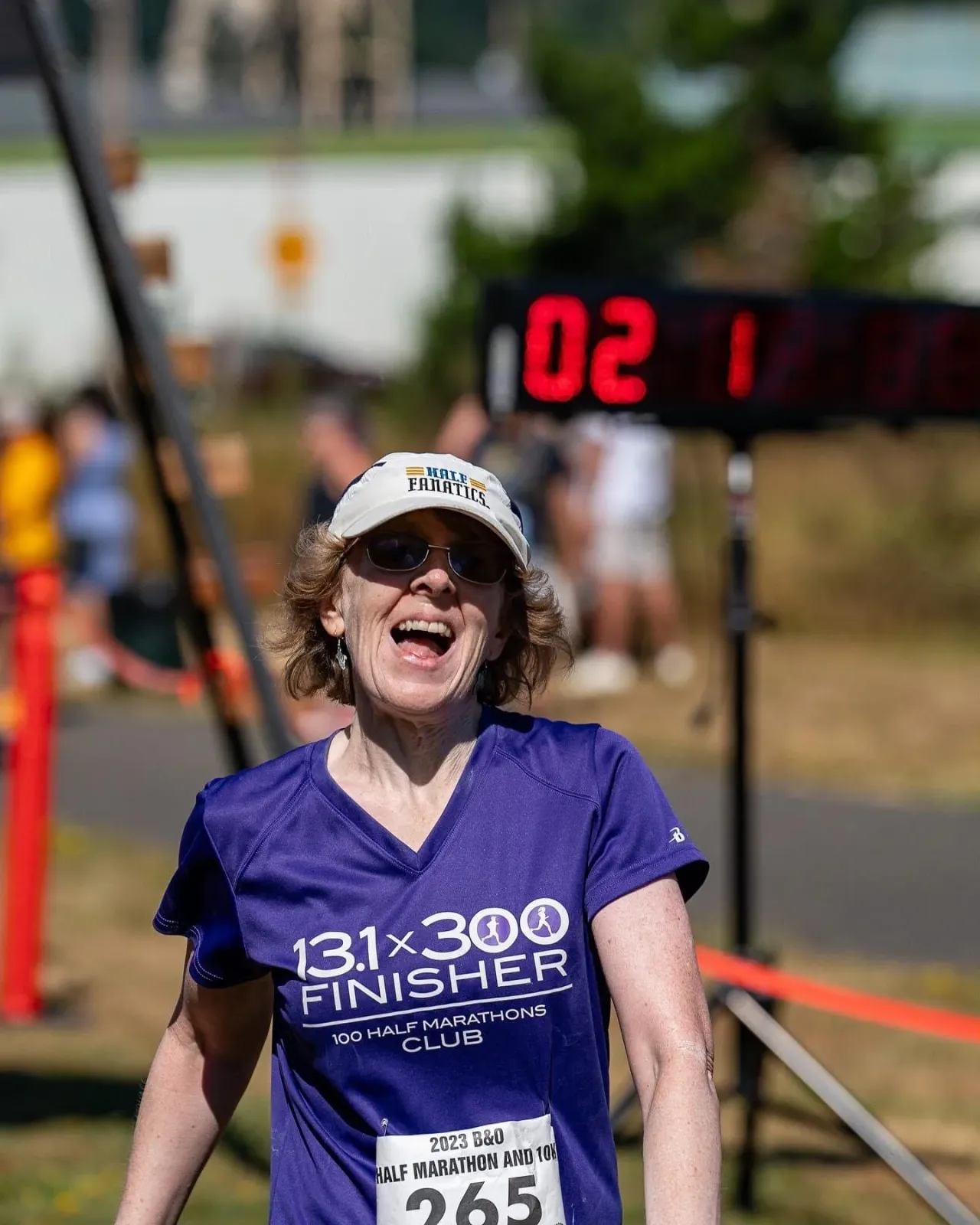 A female runner with an ecstatic expression crosses the finish line, wearing a purple shirt and a white cap.