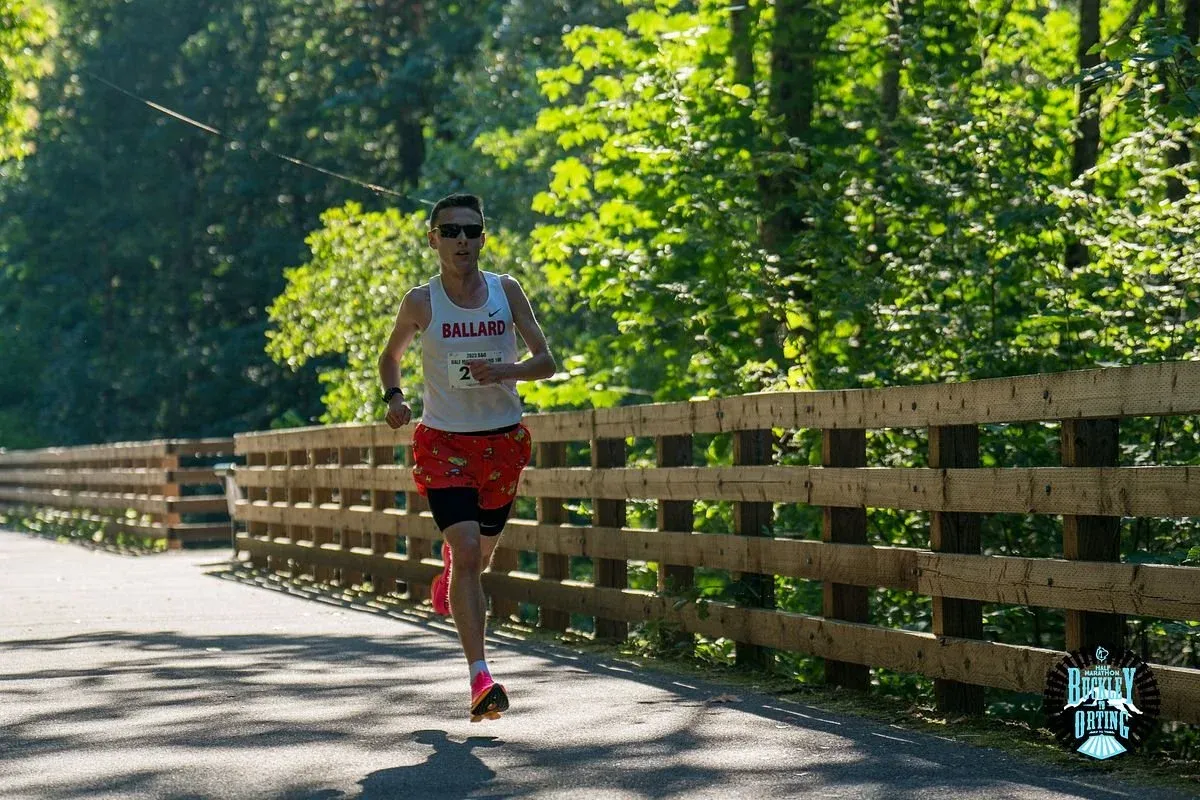 A male runner with sunglasses and a white tank top with 'BALLARD' and the number '2' on it, runs on a paved path next to a wooden fence with trees in the background.