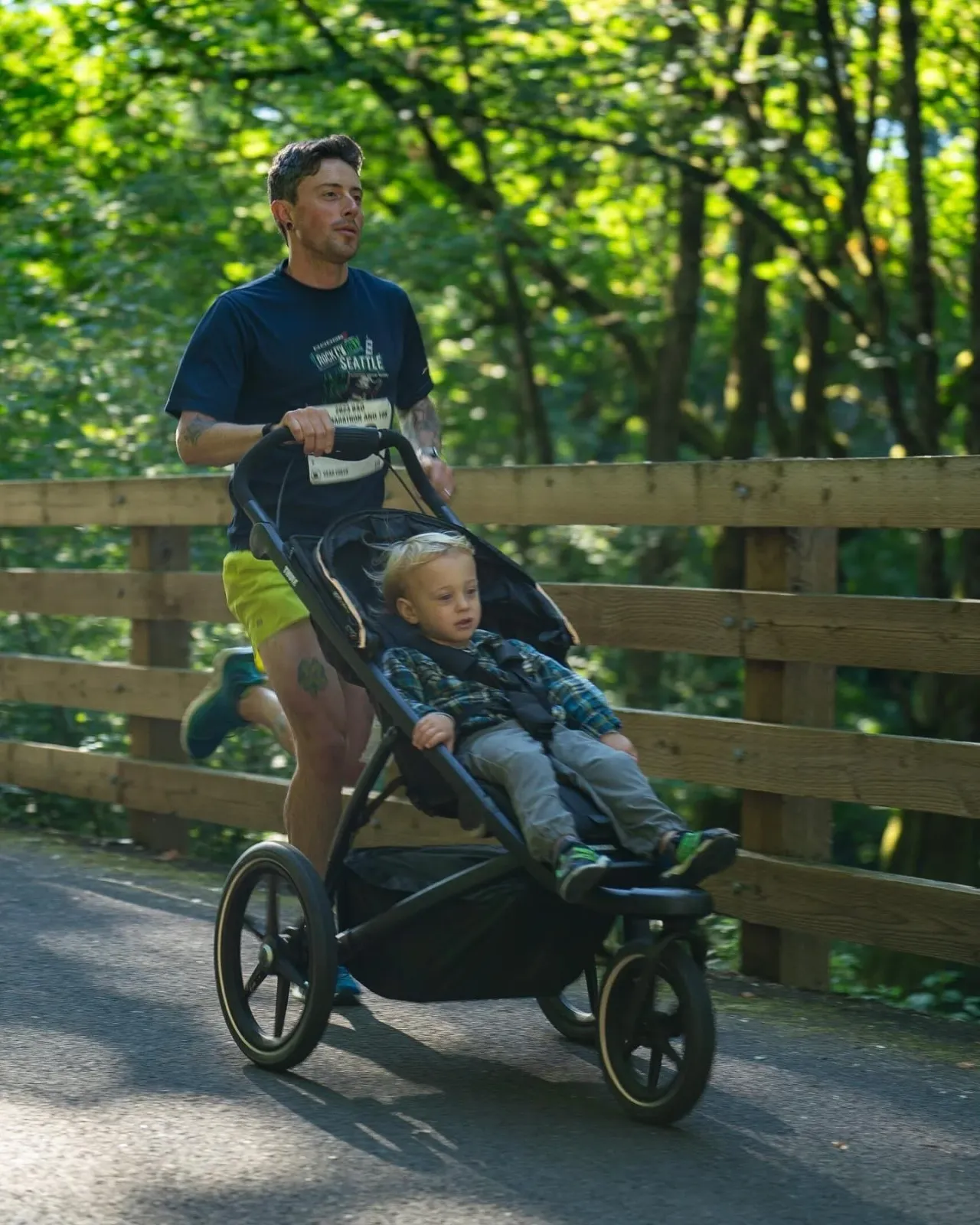 A man runs on a paved path pushing a stroller with a young child in it, with a wooden fence and trees in the background.