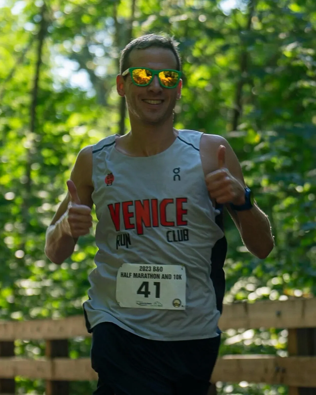Runner with bib number 41 and 'Venice Run Club' shirt gives a thumbs up while running on a wooded path.