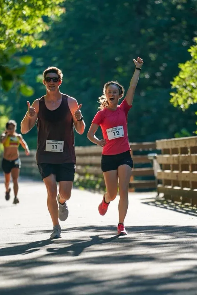 A male runner gives a thumbs up while running alongside a female runner who has her arm raised in celebration, with another runner in the background.