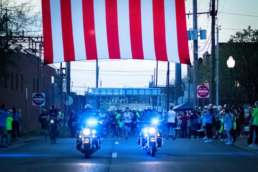 Runners at the start line of a race, with police motorcycles in the foreground and a large red and white banner overhead.