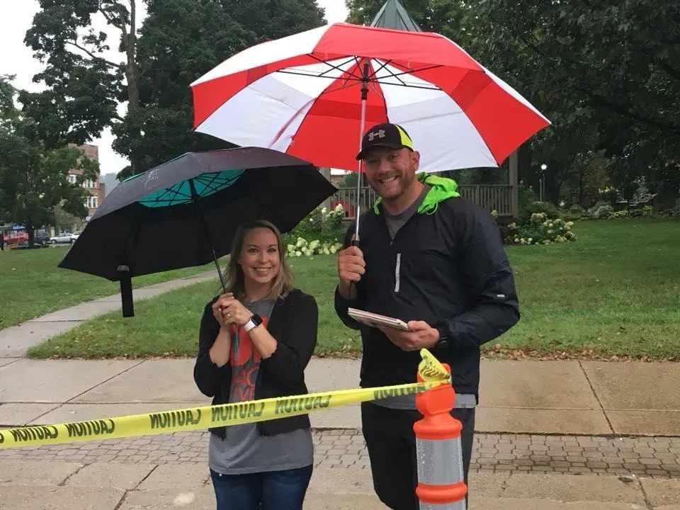 A man and a woman smiling and holding umbrellas, with caution tape in the foreground.