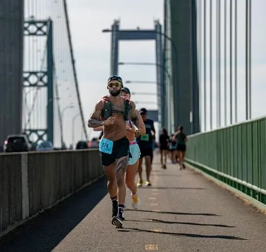 A runner with bib number 174 runs across a bridge during a race, with other runners and a large suspension bridge in the background.