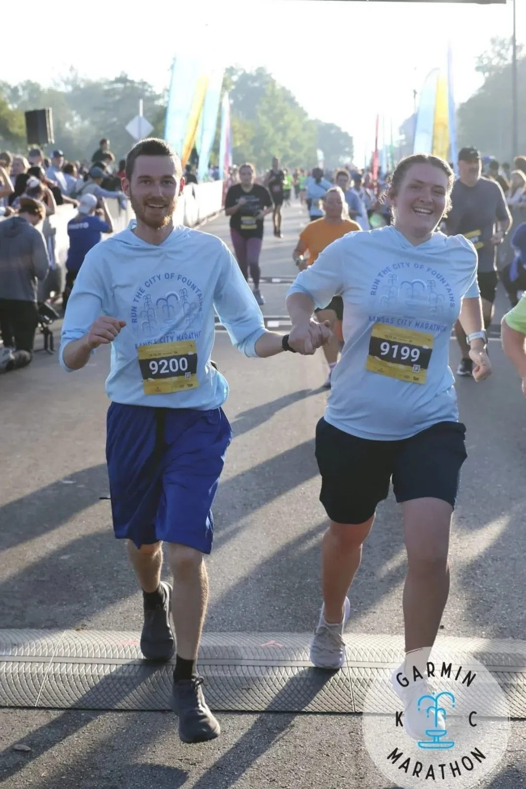 A smiling couple holds hands as they run across the start line of the Garmin Kansas City Marathon, with other runners and banners in the background.