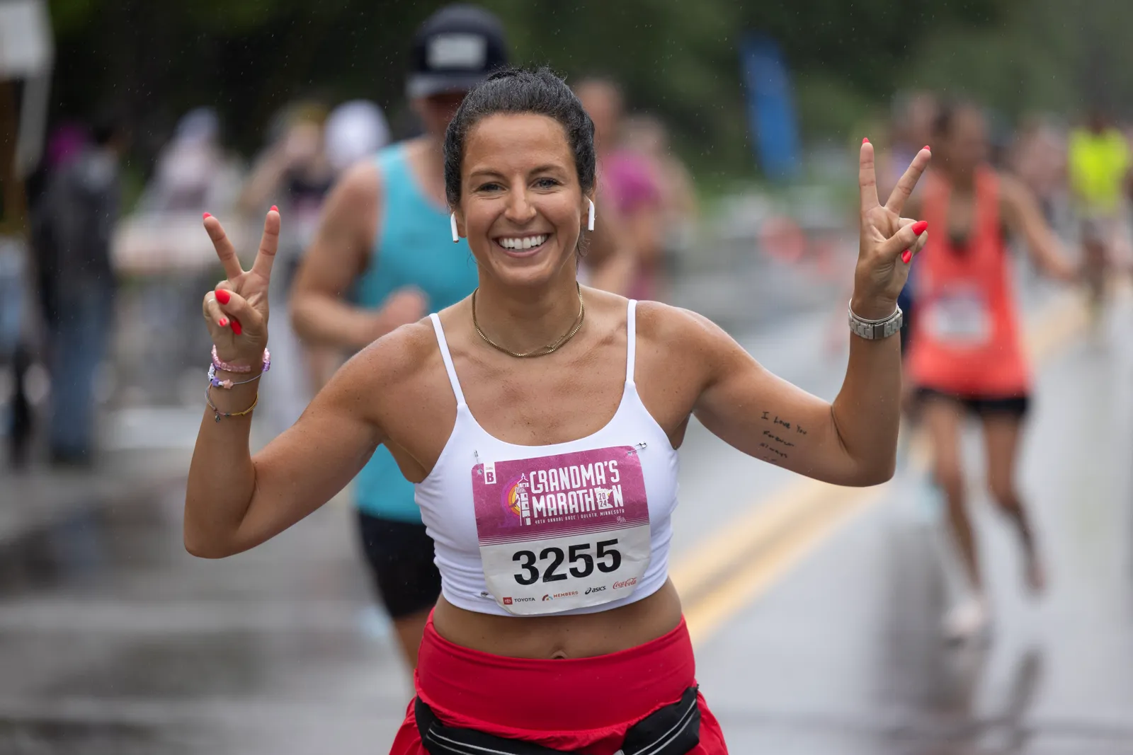 A female runner smiles and holds up two fingers in a peace sign while running in a race, with other runners blurred in the background.