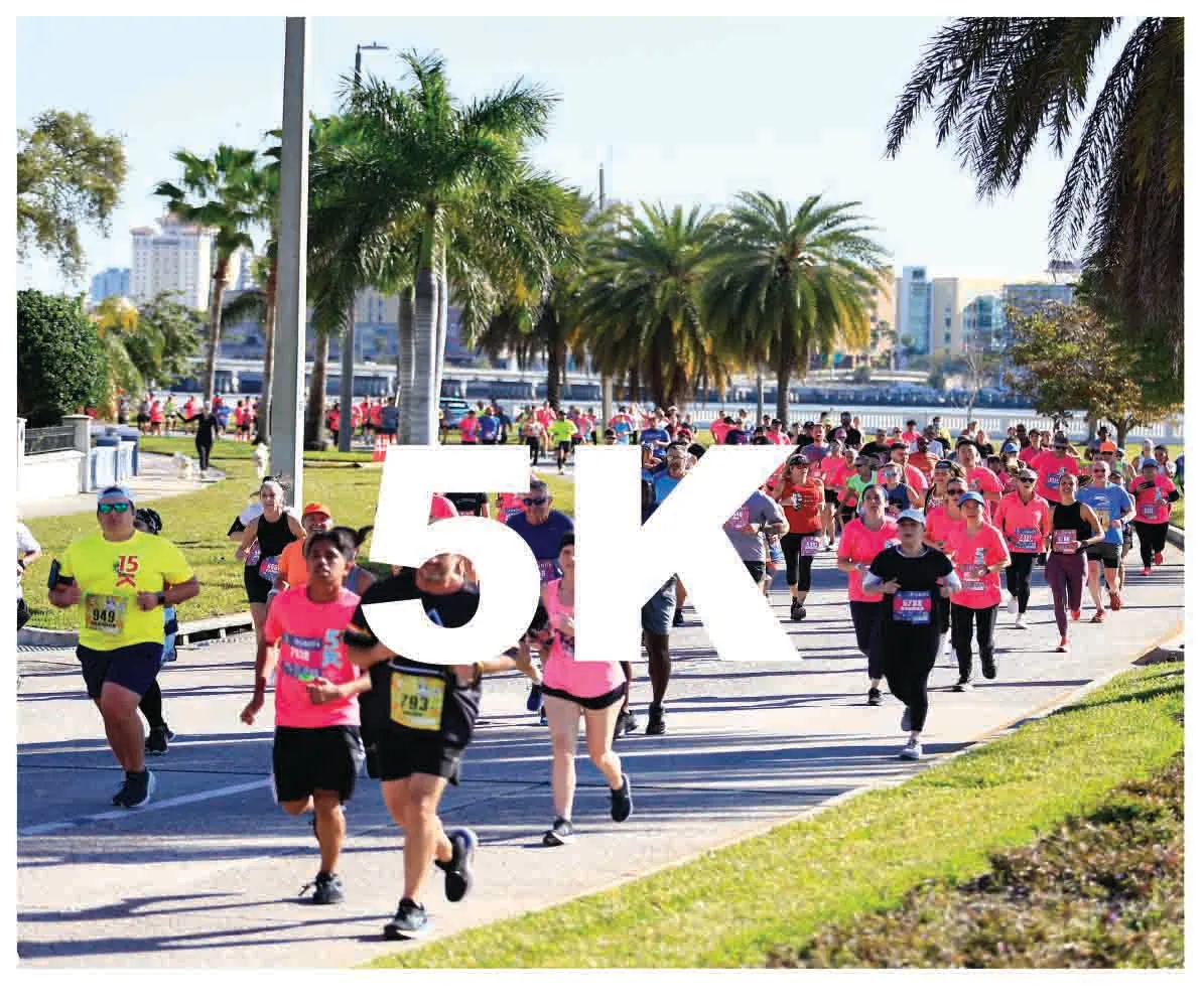 A large group of runners, mostly women in pink shirts, race down a sunny road lined with palm trees. The number '5K' is overlaid in large white letters across the image.