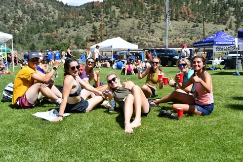 A group of young adults sitting on the grass, smiling and holding red cups, with a scenic mountain backdrop.