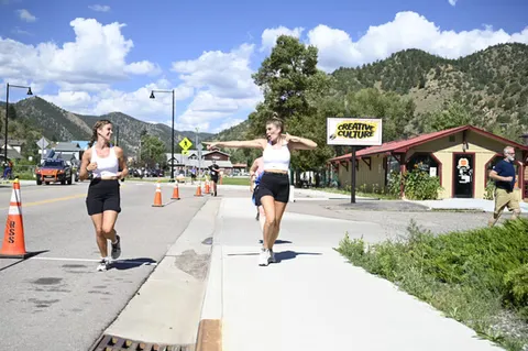 Two female runners smiling as they run on a road with mountains in the background.