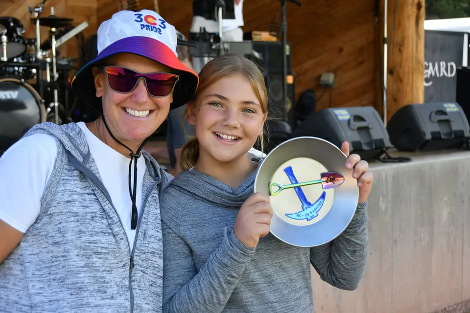 A woman and a young girl smile at the camera, holding a medal shaped like a gold pan with a pickaxe and shovel.