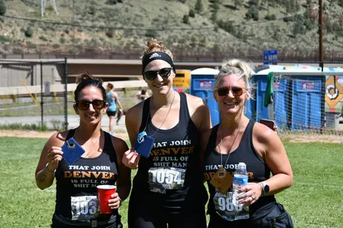 Three women in black tank tops, smiling and holding race bibs, with portable toilets and a hilly background.