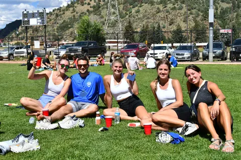 A group of five smiling people sitting on the grass, holding red cups, with a scenic mountain backdrop.
