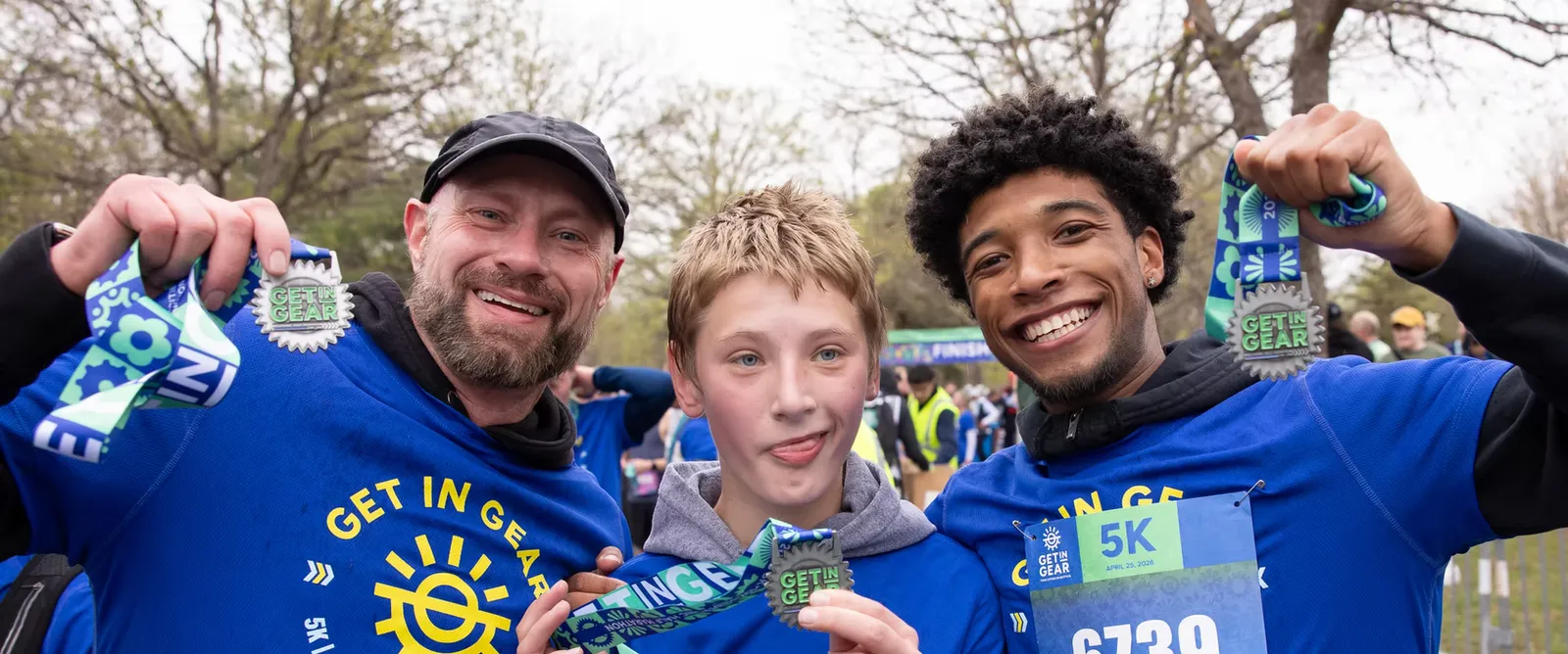 Three smiling people, two adults and a child, proudly display their race medals after completing the Get In Gear 5K race.
