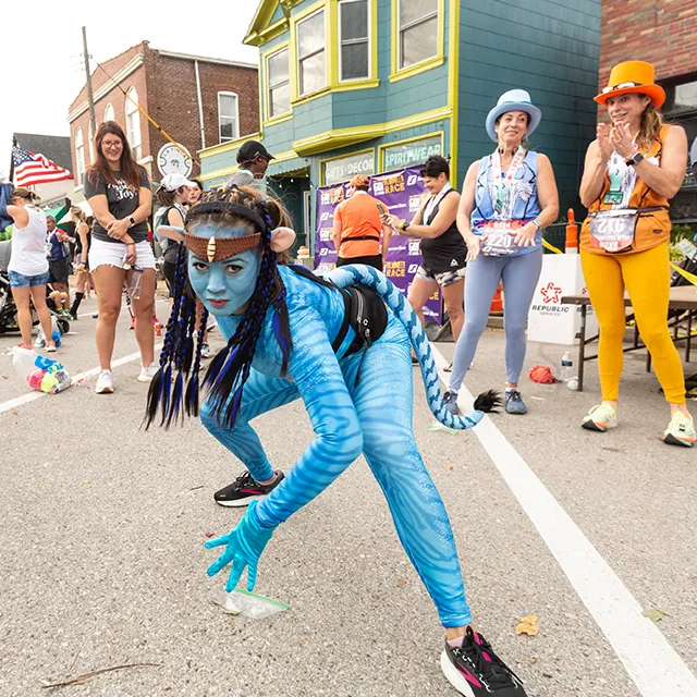 A runner dressed as a character from Avatar poses at a Halloween-themed race, with other participants and spectators in the background.