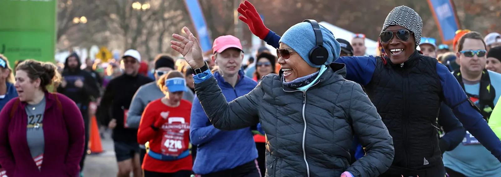A diverse group of runners, some smiling and waving, are at the start of a race, with a green banner and blue arch in the background.