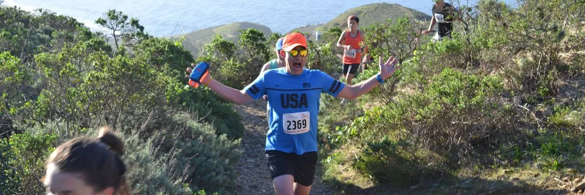 A runner in a blue 'USA' shirt and orange hat celebrates with arms outstretched while running on a scenic trail overlooking the ocean.